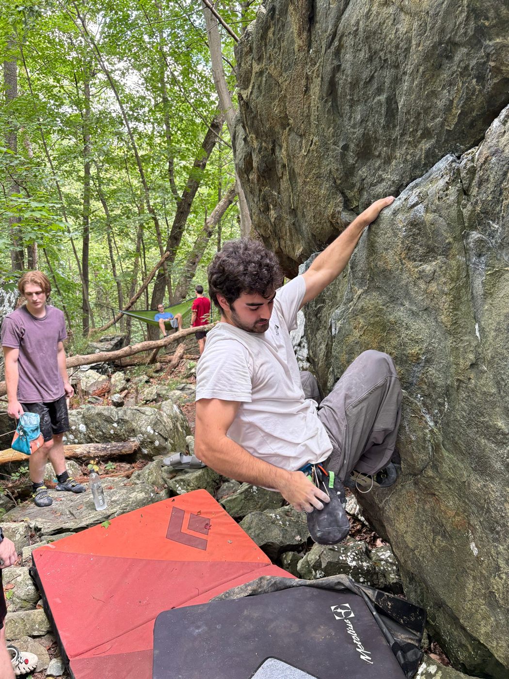 Gabe reaching high on a steep boulder in summer woods, crash pads and a spotter below
