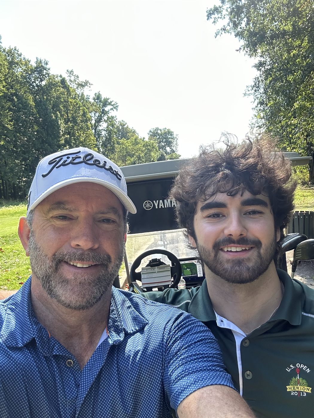 Gabe and his dad in a golf cart on a wooded course on a bright day